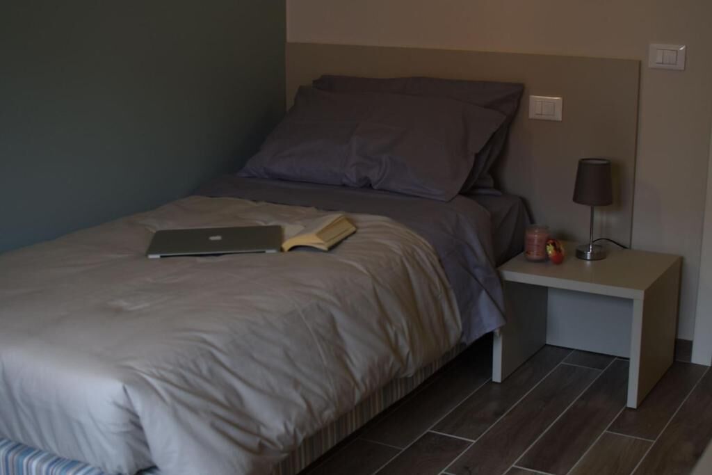 Un lit bien fait, avec un ordinateur portable et un livre, une table de chevet avec une lampe, et une petite table avec des fruits.