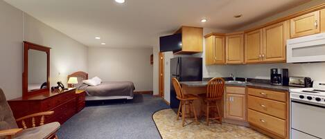 A compact bedroom with a single bed, a wooden dresser, and a mirror. Adjacent is a kitchenette with a stove, microwave, and refrigerator.