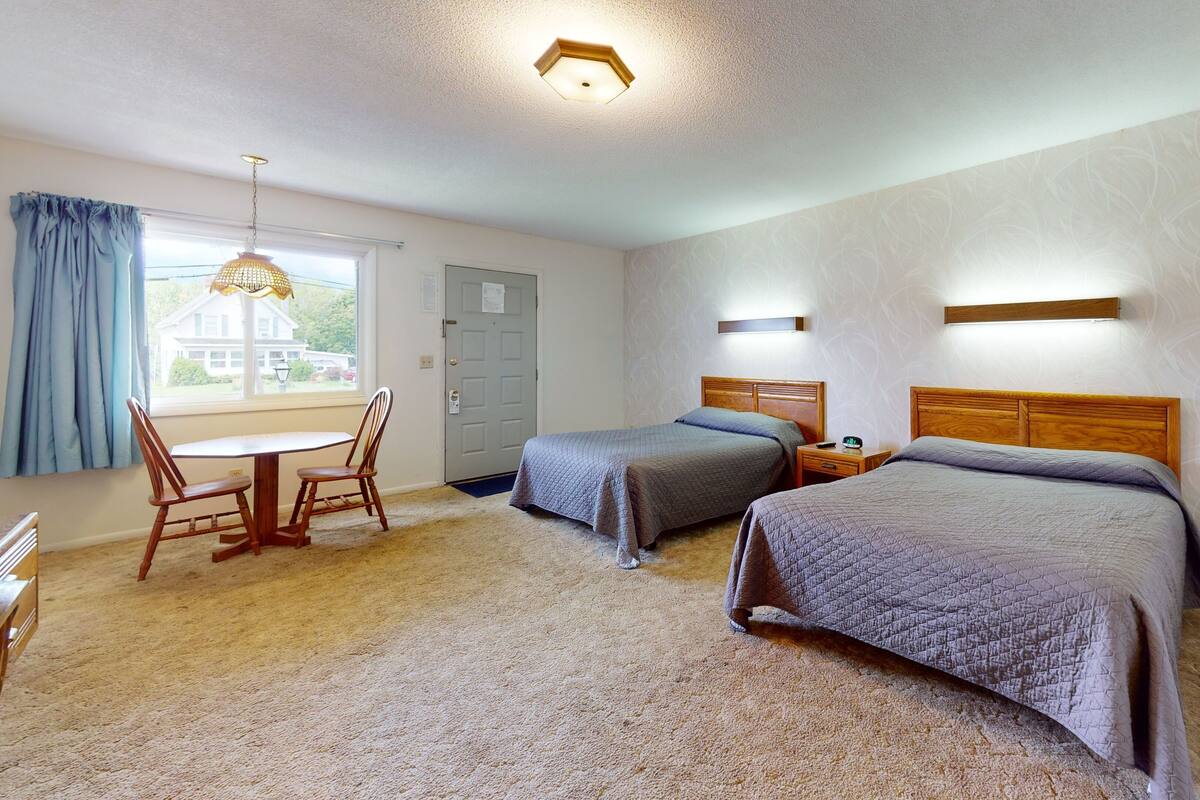 Habitación de hotel con dos camas, un cabecero de madera, una mesita con una silla, una ventana con cortinas y una puerta.