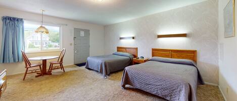 Habitación de hotel con dos camas, un cabecero de madera, una mesita con una silla, una ventana con cortinas y una puerta.