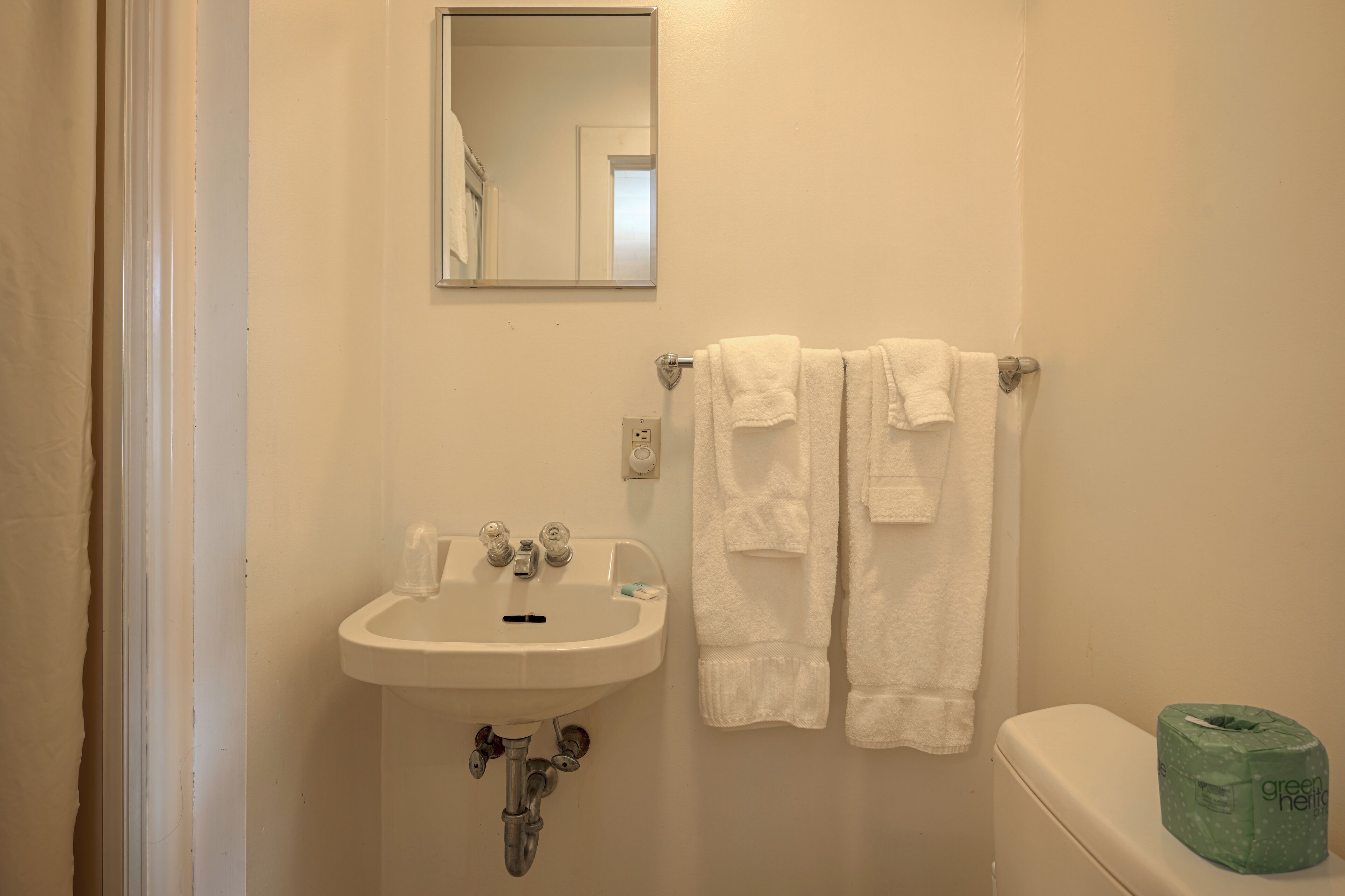 Un baño con un lavamanos blanco, un espejo y dos toallas blancas colgadas en un perchero.