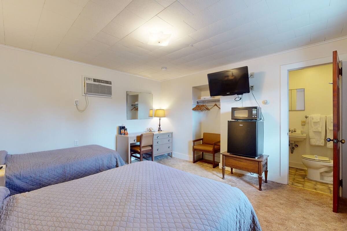 Habitación de hotel con dos camas, un escritorio con silla, televisor montado en la pared, microondas y baño visible a través de una puerta abierta.