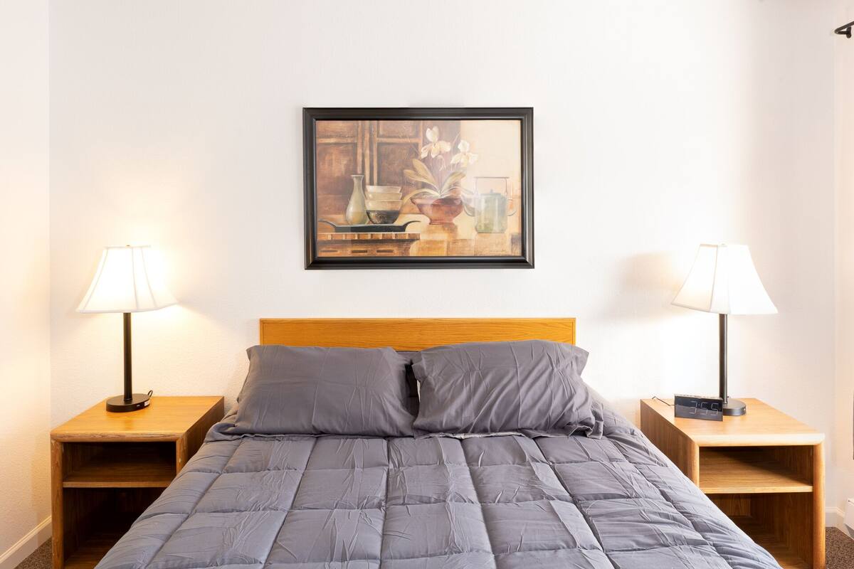 Ein ordentlich bezogenes Bett mit einer grauen Bettdecke, an beiden Seiten zwei Nachttische mit Lampen, und ein gerahmtes Bild darüber.