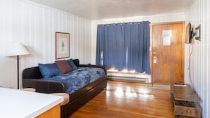 A compact living space with a sofa bed, a small table, a lamp, a framed picture, a wooden door, and a window with curtains. - Siesta Motel (Colfax)