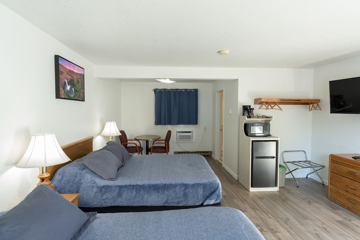 Ein Hotelzimmer mit zwei Betten, einem Schreibtisch mit Computer, einer Mikrowelle und einem kleinen Kühlschrank. In der Ecke befindet sich ein Sessel und ein Tisch, und an der Wand ist ein Fernseher angebracht.