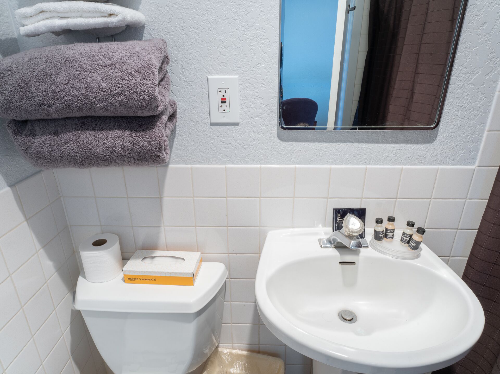 Ein Badezimmer mit einem weißen Waschbecken, einer Toilette und einem Spiegel. Handtücher sind an der Wand gestapelt, und auf der Toilette liegt eine Packung Toilettenpapier.