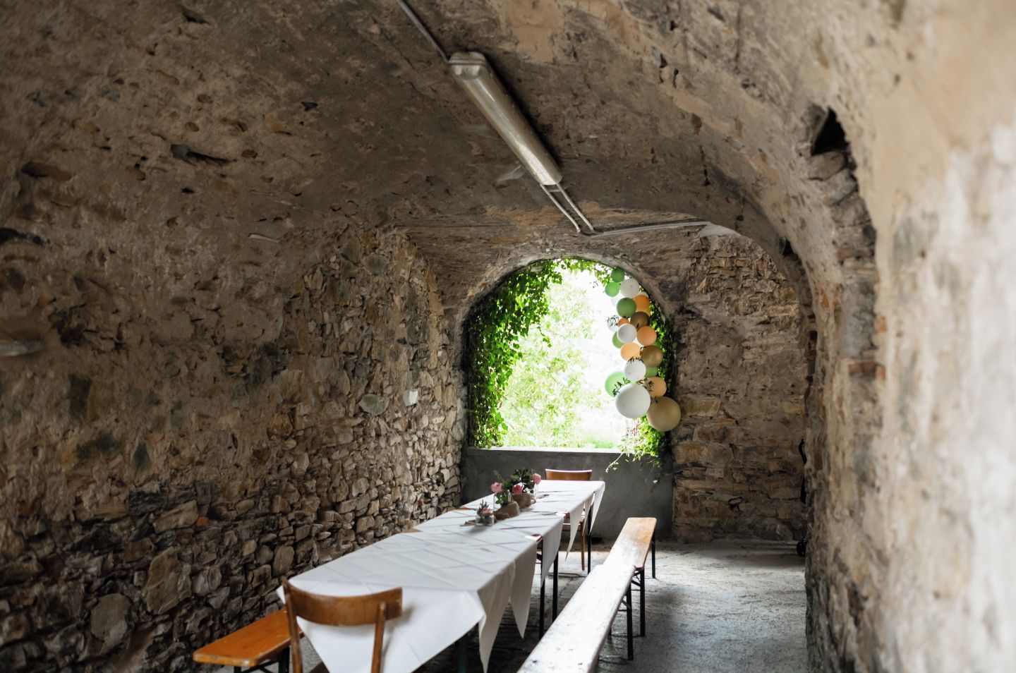 An arched stone room with a long table set for dining, a bench, and a view through the arch to greenery outside.