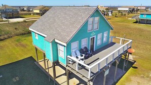 A turquoise house with a balcony, elevated on stilts, featuring a small porch with outdoor seating.