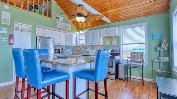 A spacious kitchen with a wooden ceiling, a central island with bar stools, and a view of the backyard through large windows.