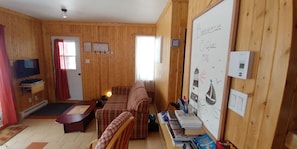 A cozy room with a wooden interior, a sofa, a television, a whiteboard with drawings, and a small table with books and a lamp. - Chalet (Saint Jean Port Joli)