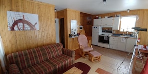 A cozy living room with a plaid sofa, a small wooden table, and a kitchen area with white cabinets and appliances. - Chalet (Saint Jean Port Joli)