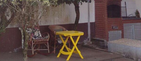 A yellow folding table and a wicker chair outside a building with a brick wall and a window.