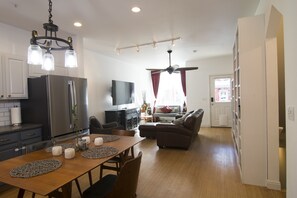 A modern kitchen with a dining area, a living room with a sofa and TV, and a hallway leading to a door.