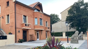 Um edifício de dois andares com fachada bege e telhado de telhas vermelhas. Possui duas varandas com grades, um conjunto de degraus que levam à entrada e uma pequena área de jardim com diversas plantas e flores.