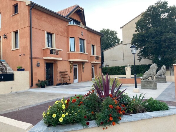 Un edificio de dos pisos con fachada color beige y tejado de tejas rojas. Cuenta con dos balcones con barandillas, una escalera que lleva a la entrada y un pequeño jardín con diversas plantas y flores.
