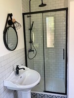 Un baño con un lavabo blanco, un espejo redondo y una zona de ducha con mampara de vidrio y marco negro.