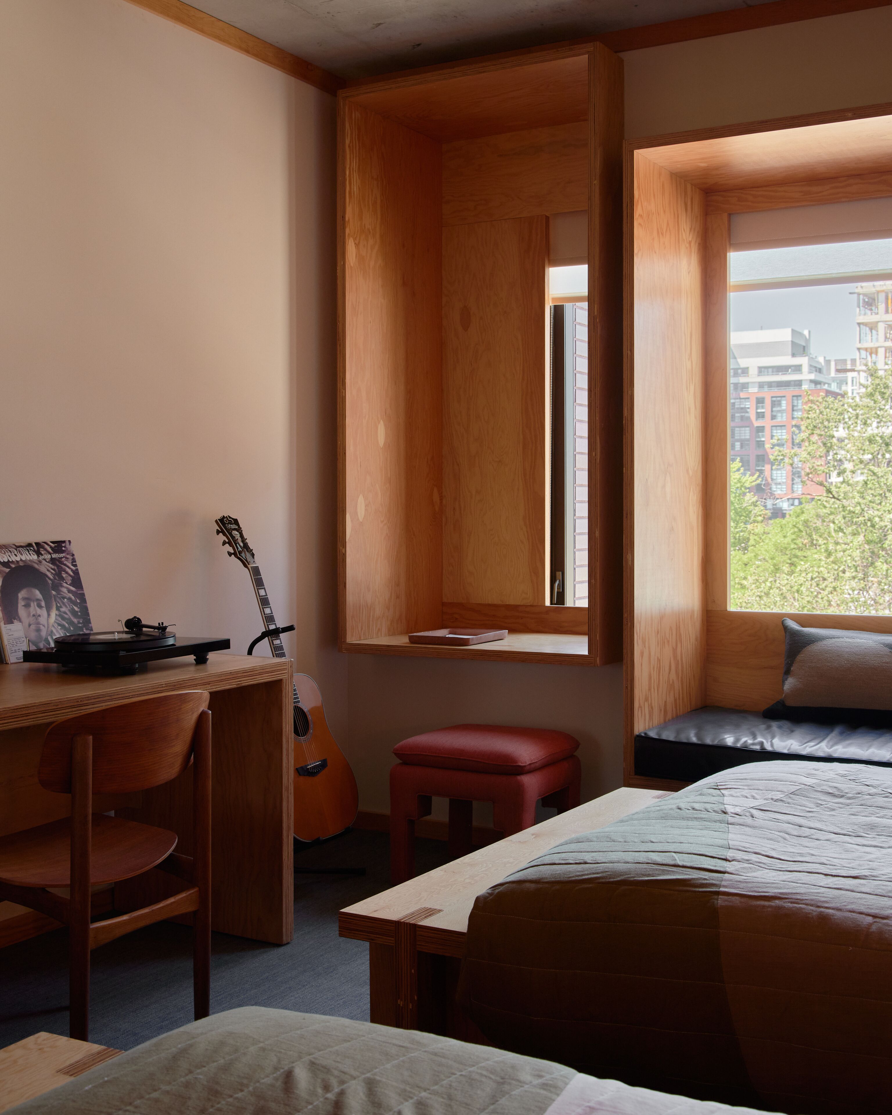 Een kamer met een houten bureau, een gitaar, een rode kruk, een bed met een kussen, en een raam met uitzicht op gebouwen.