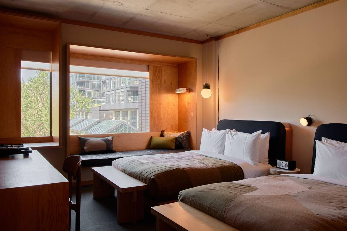 Habitación de hotel con dos camas, un escritorio y un banco. Se ve una ventana con vista a edificios.
