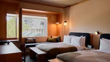 Habitación de hotel con dos camas, un escritorio y un banco. Se ve una ventana con vista a edificios.