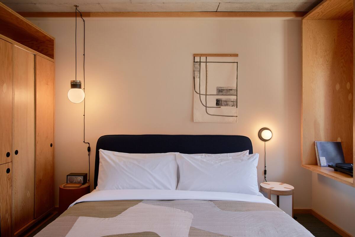 Una habitación de hotel ordenada con una cama, mesitas de noche, un armario y una obra de arte colgada en la pared.