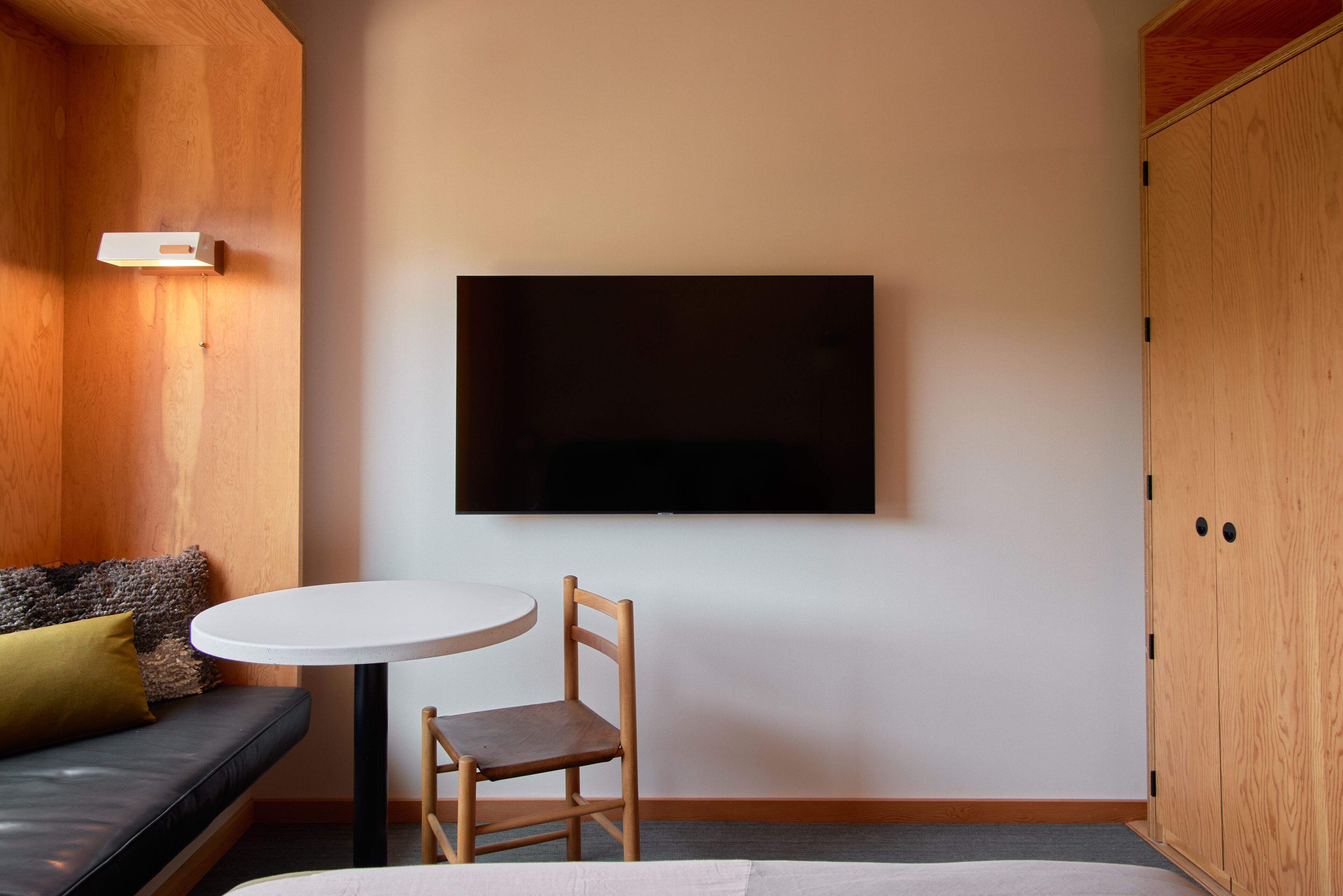 Una habitación con un armario de madera, una mesa redonda blanca con una silla, un sofá negro con un cojín verde y un televisor de pantalla plana montado en la pared.