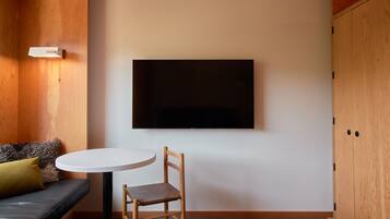 Una habitación con un armario de madera, una mesa redonda blanca con una silla, un sofá negro con un cojín verde y un televisor de pantalla plana montado en la pared.