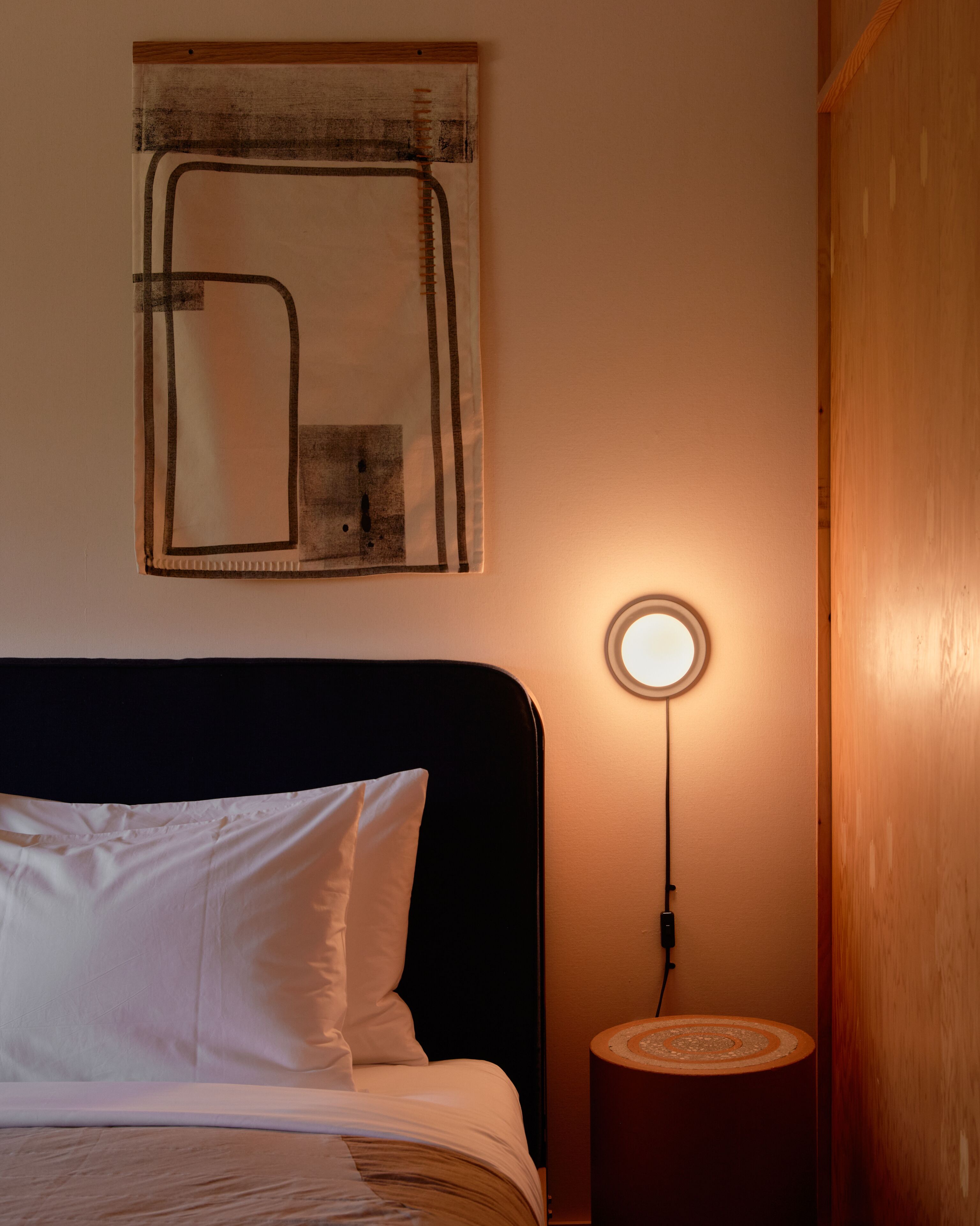 A neatly made bed with white linens, a round table lamp, and a wall-mounted abstract artwork.