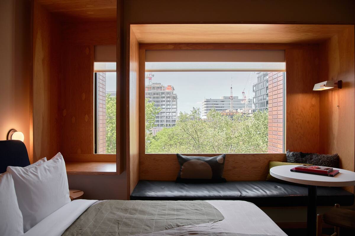 Habitación de hotel con cama, una pequeña mesa redonda, un banco con cojines y un amplio ventanal con vistas a edificios y árboles.