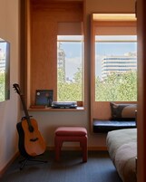 A bedroom with a guitar, a small table with a record player, a bench, and a bed with a view of buildings and trees.
