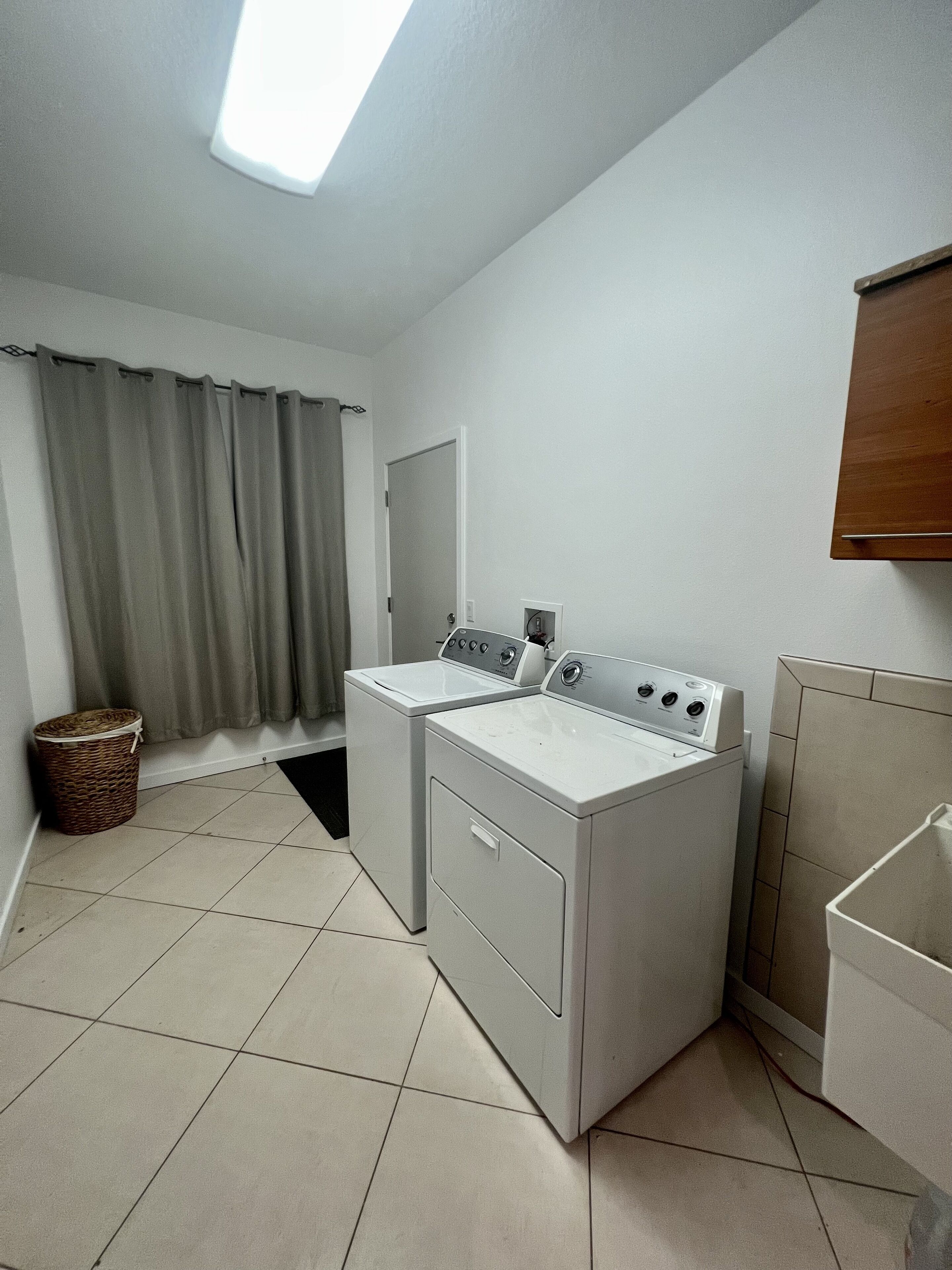A laundry room with two washing machines, a drying machine, a window with curtains, and a tiled floor.