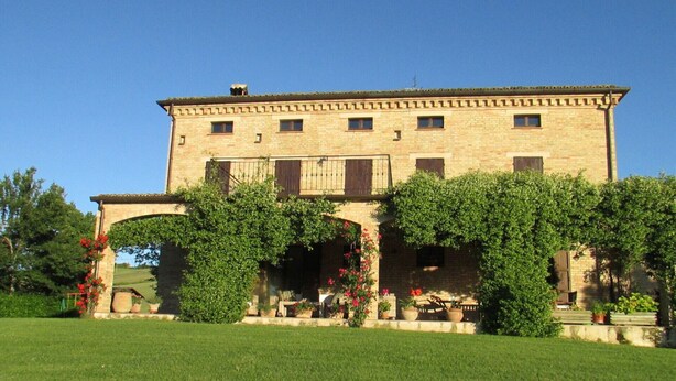 A large, two-story stone building with a central archway, surrounded by lush greenery and potted plants. - Villa Giulia - Homelike Villas (Appignano)