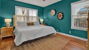 A bedroom with teal walls, a wooden floor, a bed with white bedding, two bedside lamps, and a ceiling fan. - Walk to Main St • Hot Tub • Backyard Oasis (Fredericksburg)