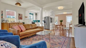 A spacious living area with a brown leather sofa, blue armchairs, and a glass coffee table. A kitchen with white cabinets and a large refrigerator is visible in the background. - Walk to Main St • Hot Tub • Backyard Oasis (Fredericksburg)