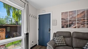 A room with a blue door, a view of trees outside, a mounted artwork, and a brown leather sofa with a plaid pillow. - The Alpenglow Loft - 1 Bed/1Bath- Radiant Charmer (Anchorage)