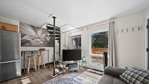 A modern kitchen with a built-in refrigerator, wooden stools, a flat-screen TV, and a staircase leading to a balcony. - The Alpenglow Loft - 1 Bed/1Bath- Radiant Charmer (Anchorage)