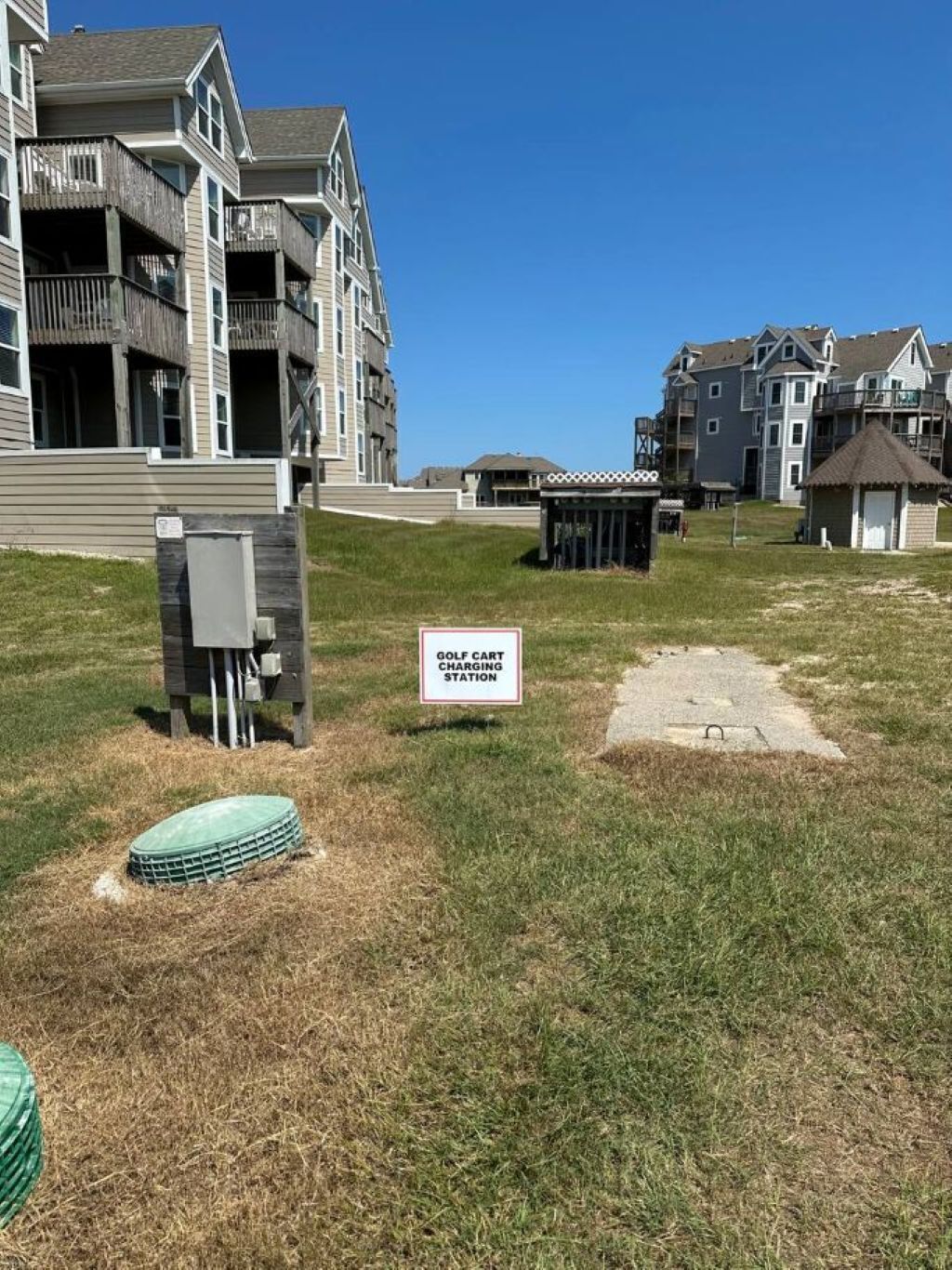 A golf cart charging station in a grassy area with multi-story residential buildings in the background.