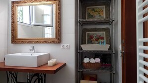 Un baño con lavamanos blanco, un espejo con marco dorado y una repisa con una cesta de toallas y un cuadro floral enmarcado.