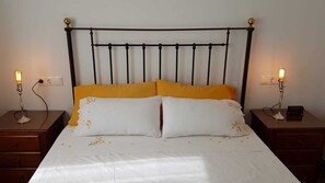 Una cama con cabecera de metal, dos mesitas de noche con lámparas y una colcha blanca con detalles amarillos.