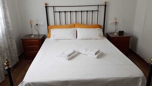 Una cama bien tendida con sábanas blancas y dos toallas enrolladas, un cabecero de metal negro y dos mesitas de noche con lámparas.