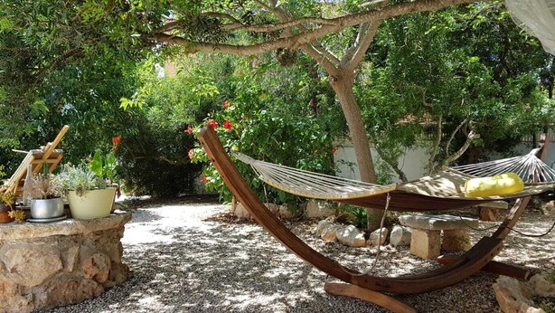 Una hamaca bajo un árbol en un jardín con plantas en macetas y un banco de piedra.