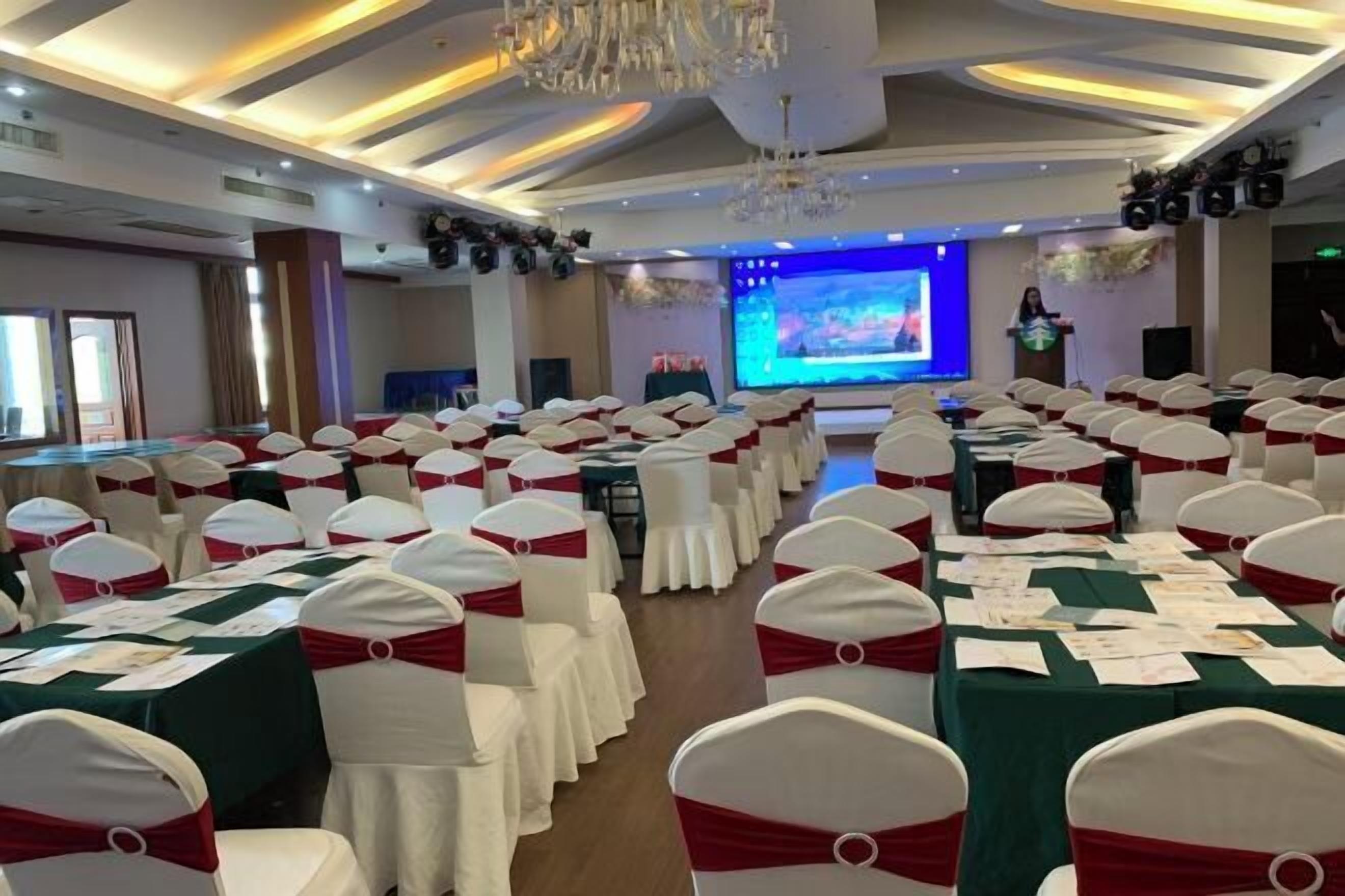 A conference room with rows of white chairs with red sashes, a stage with a large screen, and a speaker at a podium.