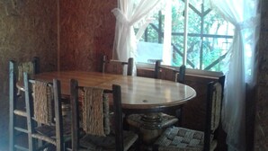 A dining area with a round wooden table and six chairs, a hanging light fixture, and a window with sheer curtains.