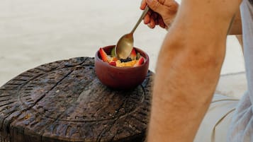 Una persona sentada en un taburete de madera en la playa, comiendo de un tazón con una cuchara.