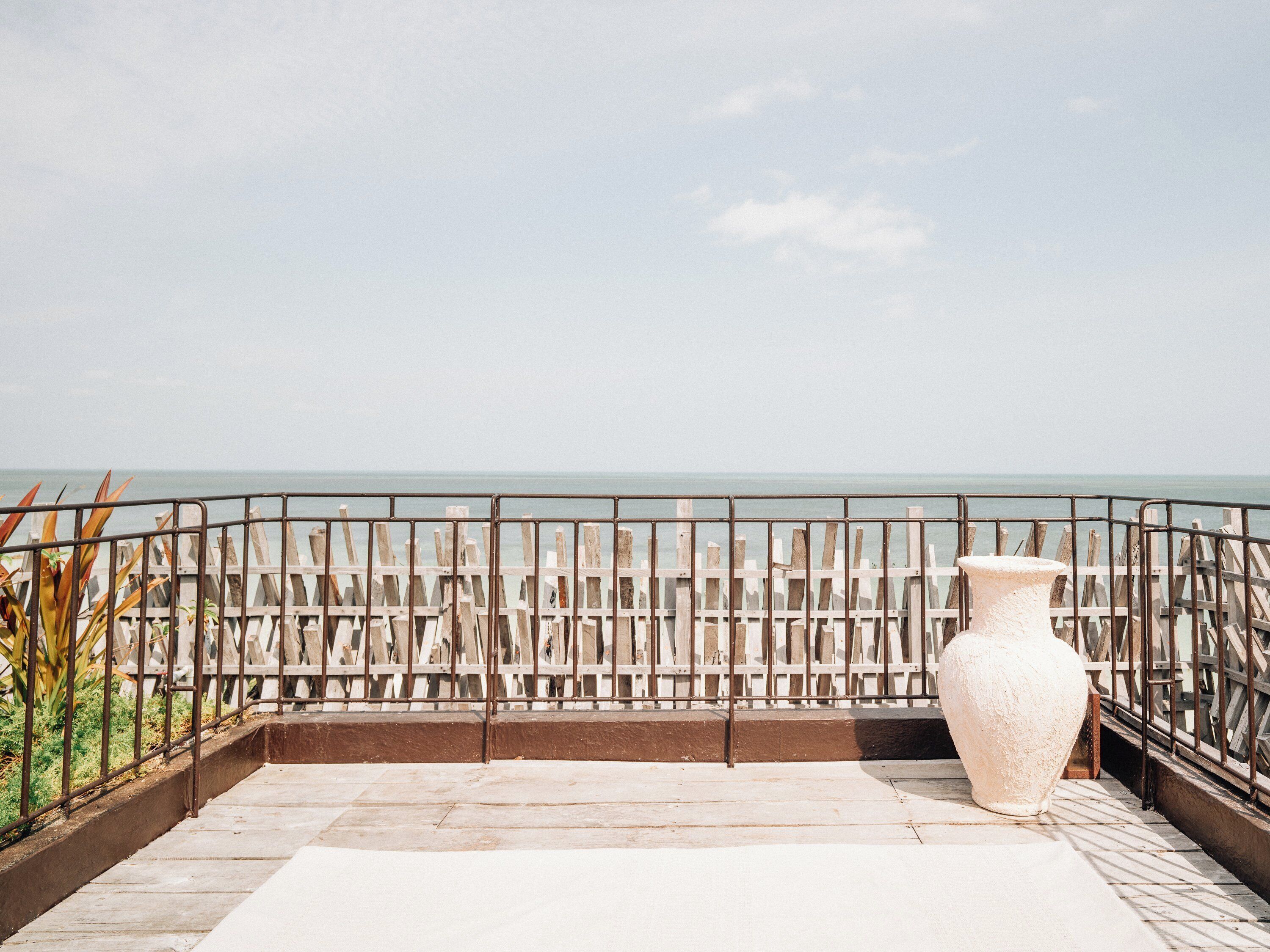 A rooftop terrace with a view of the sea, a railing, and a large white vase.