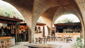 An outdoor seating area with stone arches, wooden furniture, and a bar in the background. - Nômade Temple Holbox (Isla Holbox)