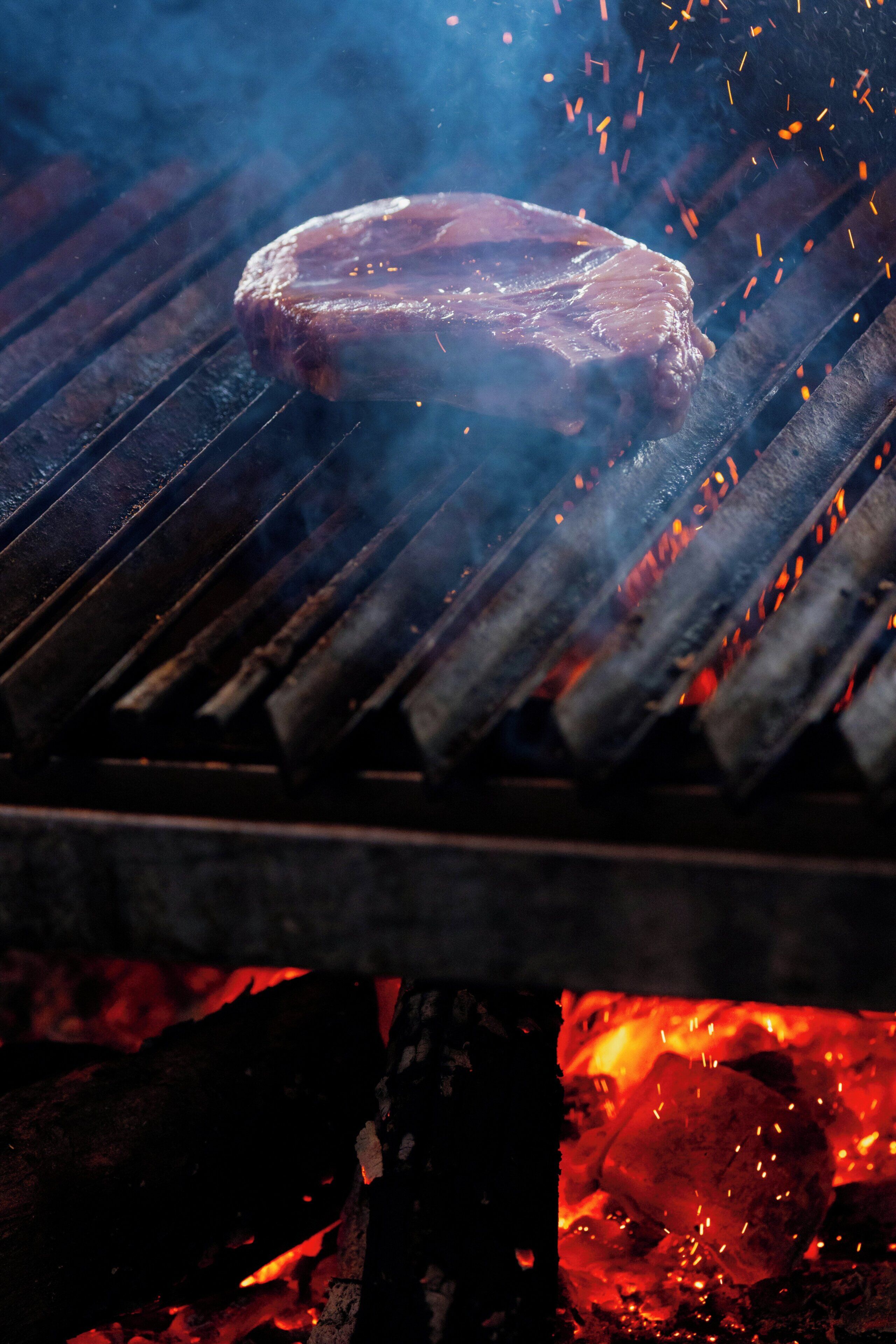 Un steak grillé sur une flamme vive, avec des braises et des étincelles visibles.