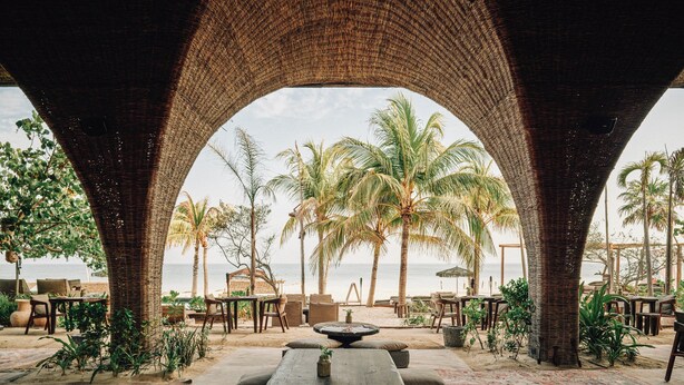 A beachside patio with wicker arches, outdoor seating, and a view of the ocean. - Nômade Temple Holbox (Isla Holbox)