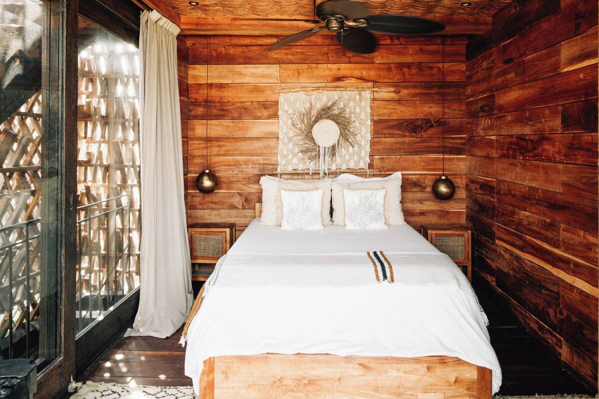 Un dormitorio de estilo cabaña de madera con una cama, un ventilador de techo y un ventanal con cortinas.