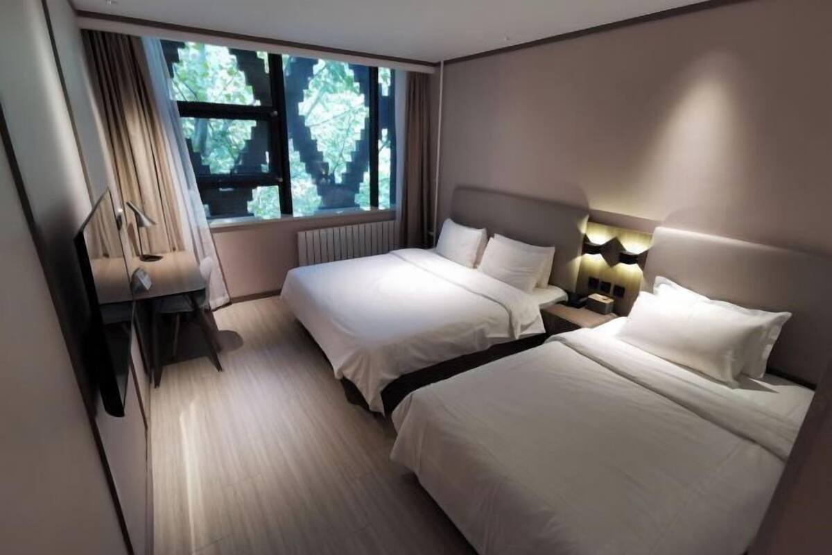 A hotel room with two beds, a desk, a chair, a lamp, and a window with a view of trees.