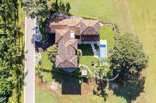 An aerial view of a house with a unique roof design, a swimming pool, and a well-maintained garden.